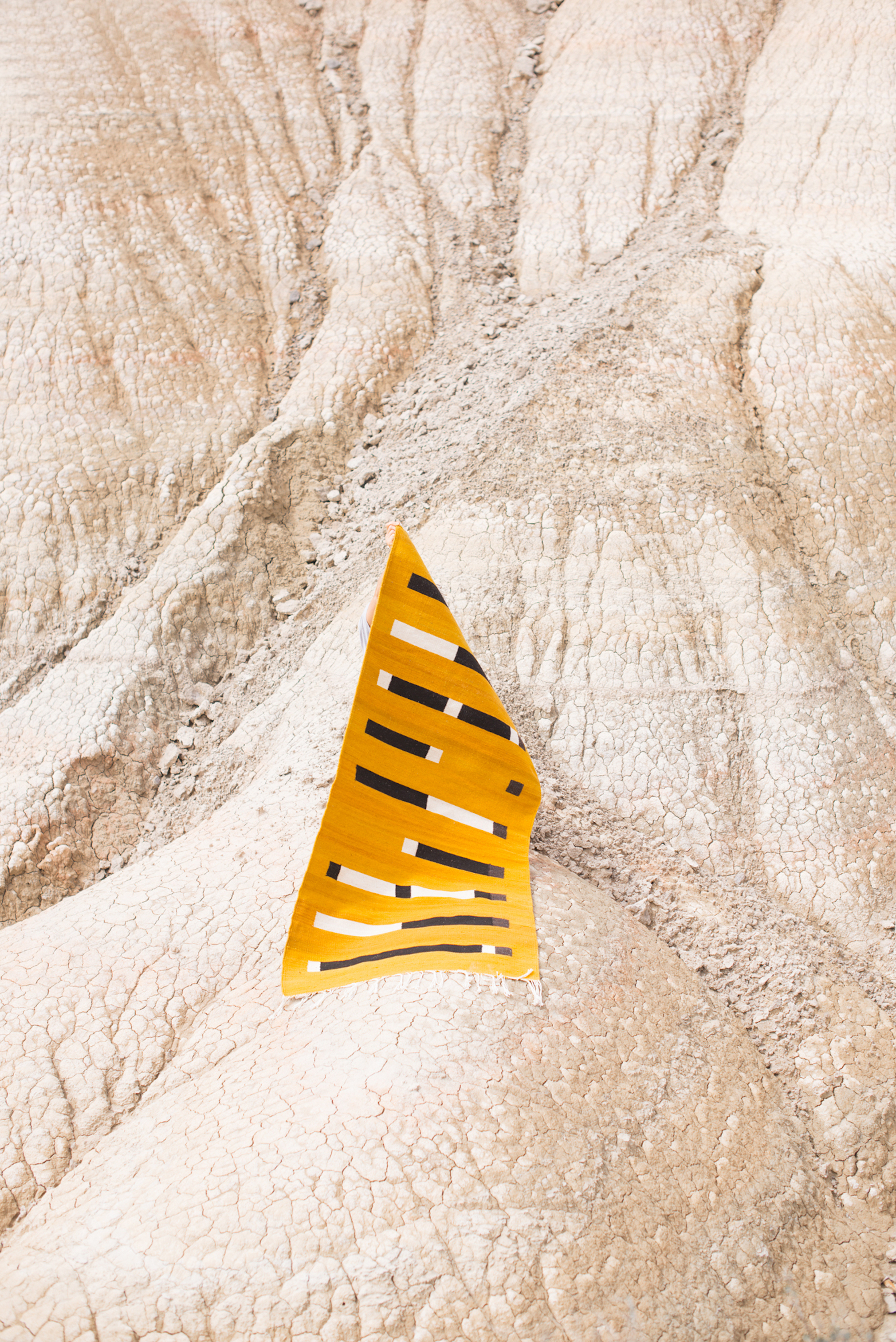 Abstract art piece featuring a yellow and white striped triangular shape against a tan, cracked earth background in a daylight outdoor shot.
