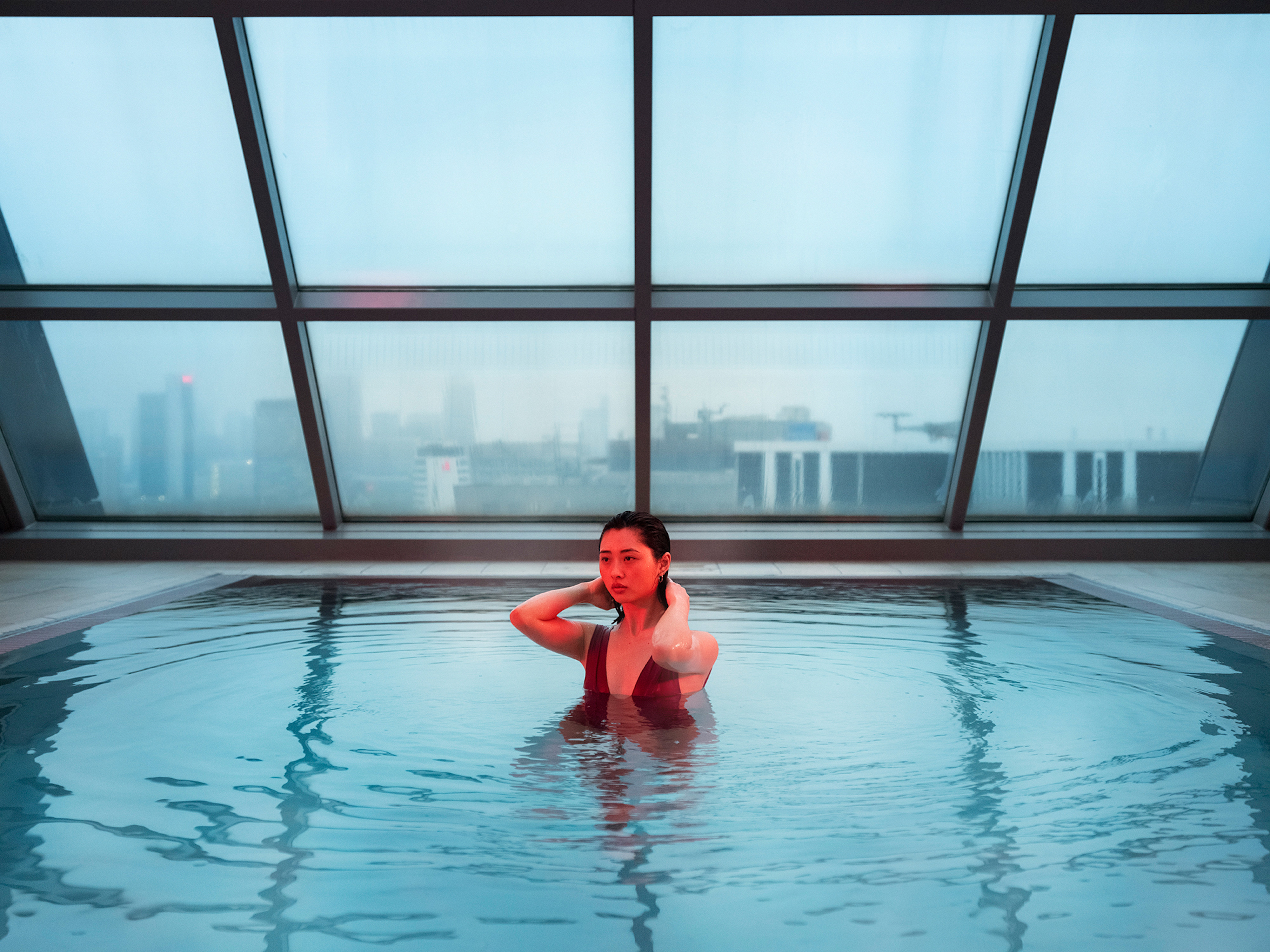 Woman in a red swimsuit in an indoor swimming pool with a city view visible through large windows, creating a serene and luxurious scene.