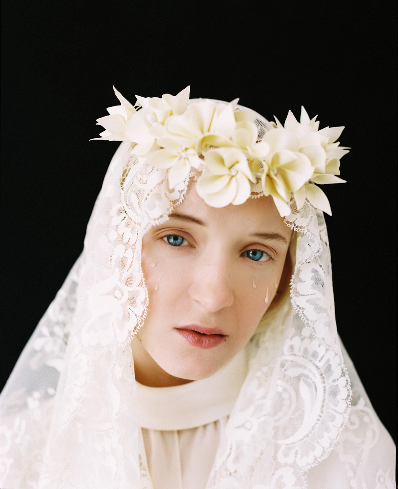 Portrait of a woman wearing a white lace veil and flower crown with visible tear drops on her cheek against a dark background