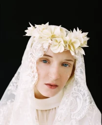 Portrait of a woman wearing a white lace veil and flower crown with visible tear drops on her cheek against a dark background