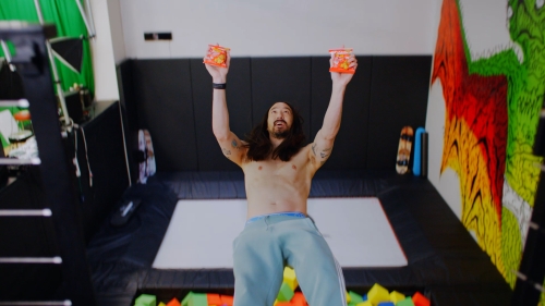 Shirtless man stands in a black-framed trampoline pit holding up two decorative pails, illuminated by bright overhead light in an indoor gym setting.