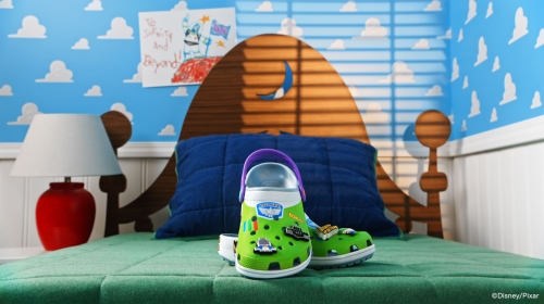 Close-up of green Crocs clogs with decorative charms on a bed with a patterned headboard, featuring colorful cartoon decor in the background.