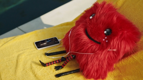 Red furry creature with a smiling face rests on a yellow towel by a pool, listening to music on a smartphone with white earbuds.