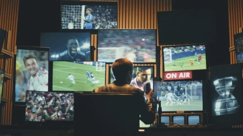 Man watches multiple screens broadcasting soccer matches in a dimly lit interior space with dark wood paneling in a media production environment.