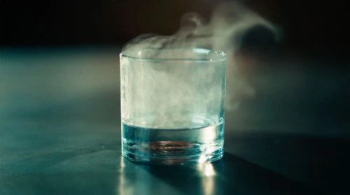 A glass of water sits on a reflective surface, with wisps of vapor or smoke rising from the liquid, against a dark background.