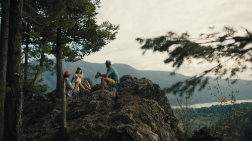 Three people, including two women and a man, sit on a rocky outcrop surrounded by trees and mountains in an outdoor setting.
