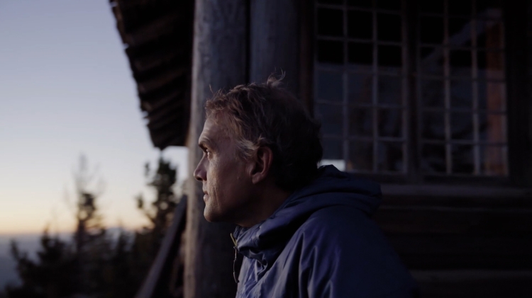 Adult man in a blue hooded jacket standing on the porch of a wooden cabin, looking toward the mountain vista at dusk.