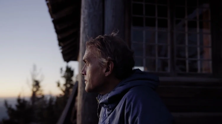 Adult man in a blue hooded jacket standing on the porch of a wooden cabin, looking toward the mountain vista at dusk.