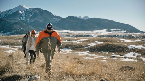 Group of four people hiking in a grassy field with snow-capped mountains in the distance, wearing outdoor clothing under a clear blue sky.