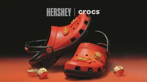 Two red Crocs with Hershey's and Reese's branding, one upright, one on its side, next to two foil-wrapped Reese's Peanut Butter Cups.