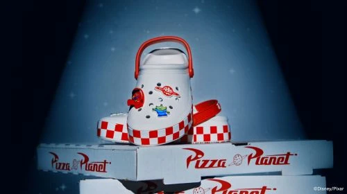 Toy Story Crocs sit atop Pizza Planet boxes, spotlighted against a dark blue backdrop with tiny white stars, showcasing a fun footwear collaboration.