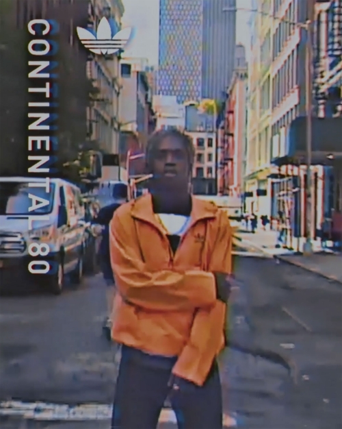 Man in orange Adidas jacket poses on city street with Continental 80 text, buildings, and car visible in background; hazy visual effects.