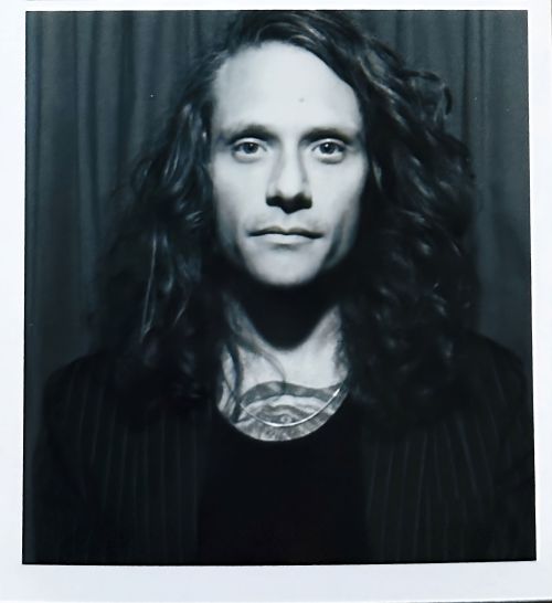 Portrait of a long-haired adult male wearing a pinstripe jacket against a dark curtain backdrop in a photo studio setting in black and white.