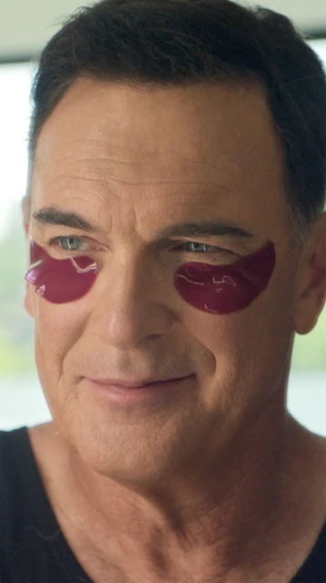 Close-up portrait of a mature man with eye patches looking at the camera, black top, neutral background, wellness and skincare concept.