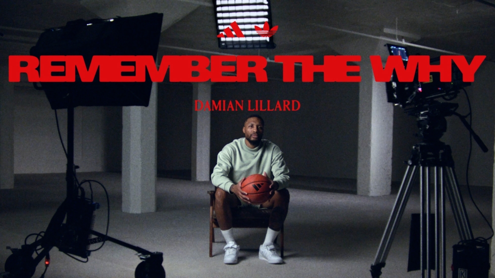 Adult male, Damian Lillard, seated, holding a basketball, centered in a promotional photo for Adidas, in a minimally furnished studio setting.