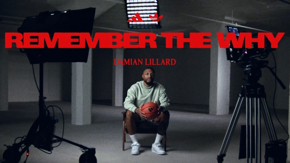 Adult male, Damian Lillard, seated, holding a basketball, centered in a promotional photo for Adidas, in a minimally furnished studio setting.
