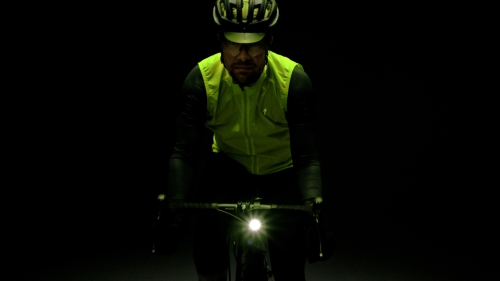 Cyclist in a helmet and vest riding a bicycle with a front headlight in a dark setting. The cyclist is looking towards the camera.