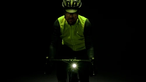 Cyclist in a helmet and vest riding a bicycle with a front headlight in a dark setting. The cyclist is looking towards the camera.