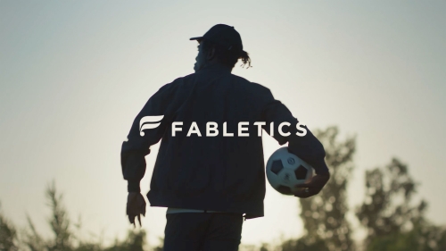 Backlit silhouette of adult male holding soccer ball, wearing athletic jacket, cap, against sky with Fabletics logo.