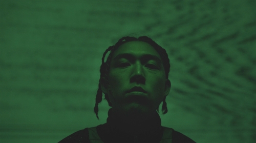 Portrait of an adult male with braided hair under green lighting, looking directly at the camera in a dimly lit indoor environment.
