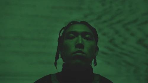 Portrait of an adult male with braided hair under green lighting, looking directly at the camera in a dimly lit indoor environment.