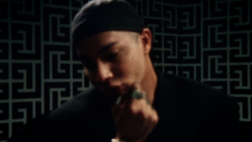 Close-up of a person in a black shirt and cap in front of a patterned wall with dark colors and shallow depth of field.
