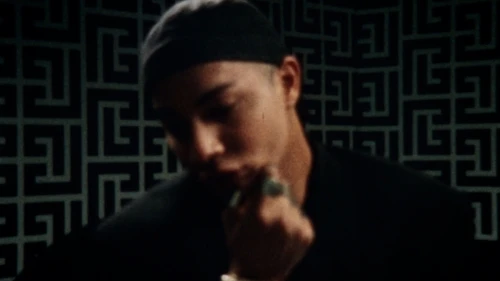 Close-up of a person in a black shirt and cap in front of a patterned wall with dark colors and shallow depth of field.