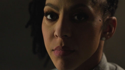 Close-up portrait of an adult female with dark hair, neutral expression, and a mole on her cheek, captured under soft, low-key lighting.