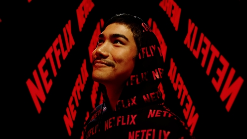 Portrait of a young adult male with a slight smile wearing a Netflix-branded hoodie against a background with the Netflix logo.