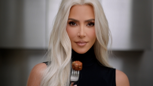 Close-up portrait of a fair-skinned adult female with long blonde hair, wearing a black turtleneck, holding a meatball on a fork.