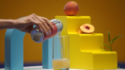 A can is being poured into a glass on a white table, with yellow steps and a whole and sliced peach in the background.