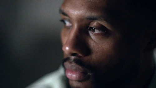 Close-up of an African American male adult looking slightly off-camera with soft lighting and neutral expression in a dark indoor space.