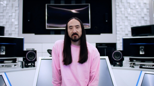 Headshot portrait of a man with long dark hair wearing a pink long-sleeve shirt in a music studio with visible audio equipment.