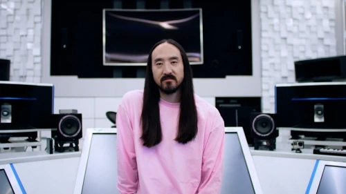 Headshot portrait of a man with long dark hair wearing a pink long-sleeve shirt in a music studio with visible audio equipment.