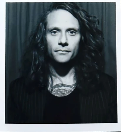 Polaroid portrait of a man with long dark wavy hair and tattoos wearing a dark jacket against dark curtains