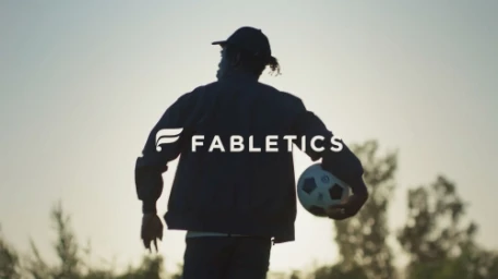Silhouette of a man wearing a cap and jacket holding a soccer ball against a bright sunset sky and trees.