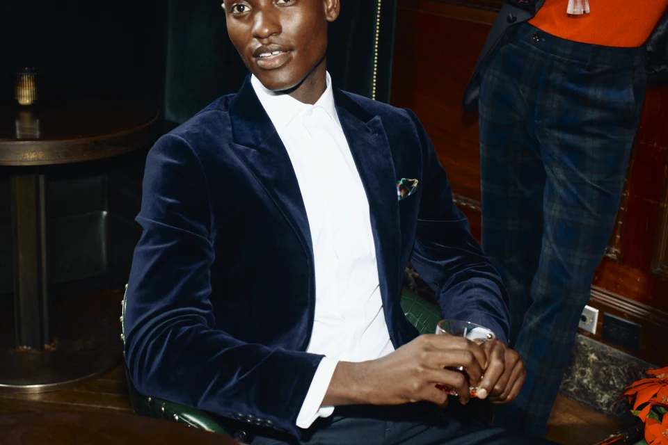 Elegant Black man in a velvet blazer and white shirt sits at a dark wood table in a bar with another person in the background.