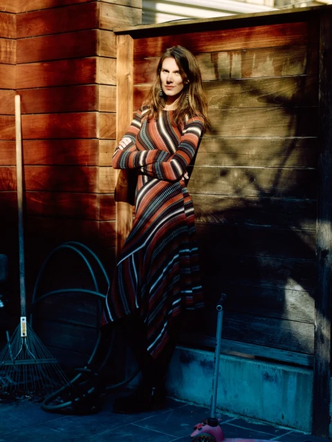 Adult woman poses with arms crossed in striped dress against wooden planks with shadows; garden tools and pink scooter visible.