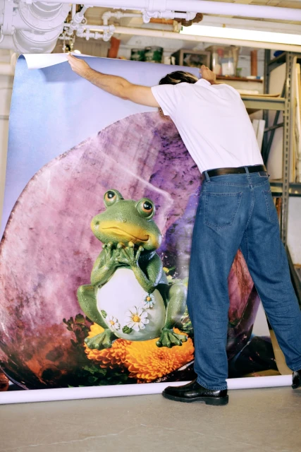 Man adjusts a large photo print featuring a whimsical frog on a flower. Image is indoors with overhead lighting and minimal decor.