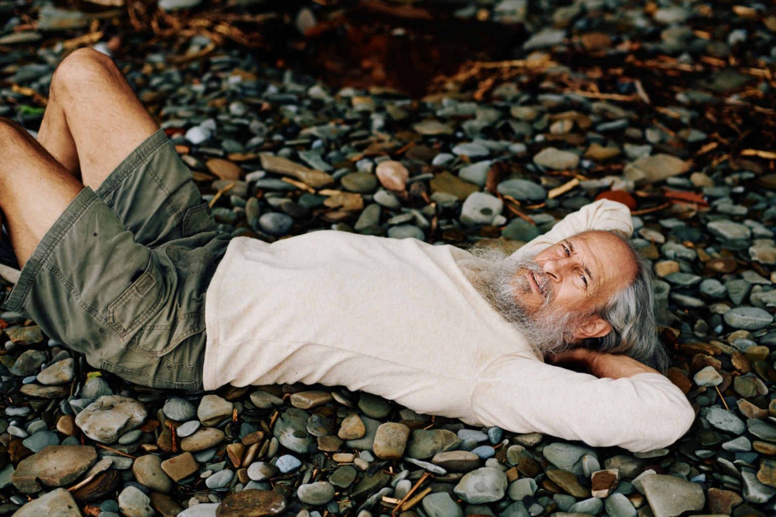 Elderly man with a beard relaxes on a rocky beach. He wears a white shirt and olive-green shorts in an outdoor, natural setting.