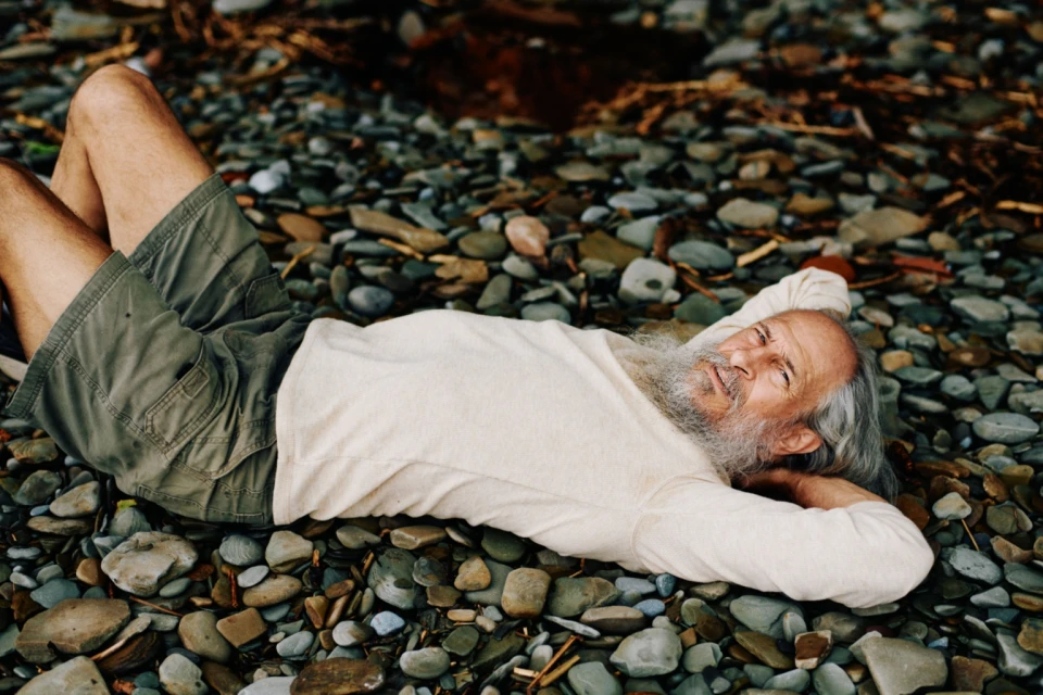 Elderly man with a beard relaxes on a rocky beach. He wears a white shirt and olive-green shorts in an outdoor, natural setting.