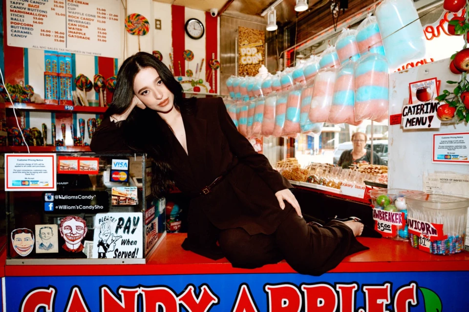 Young woman in a dark suit poses in a candy store with striped walls, surrounded by colorful sweets and vintage signage.