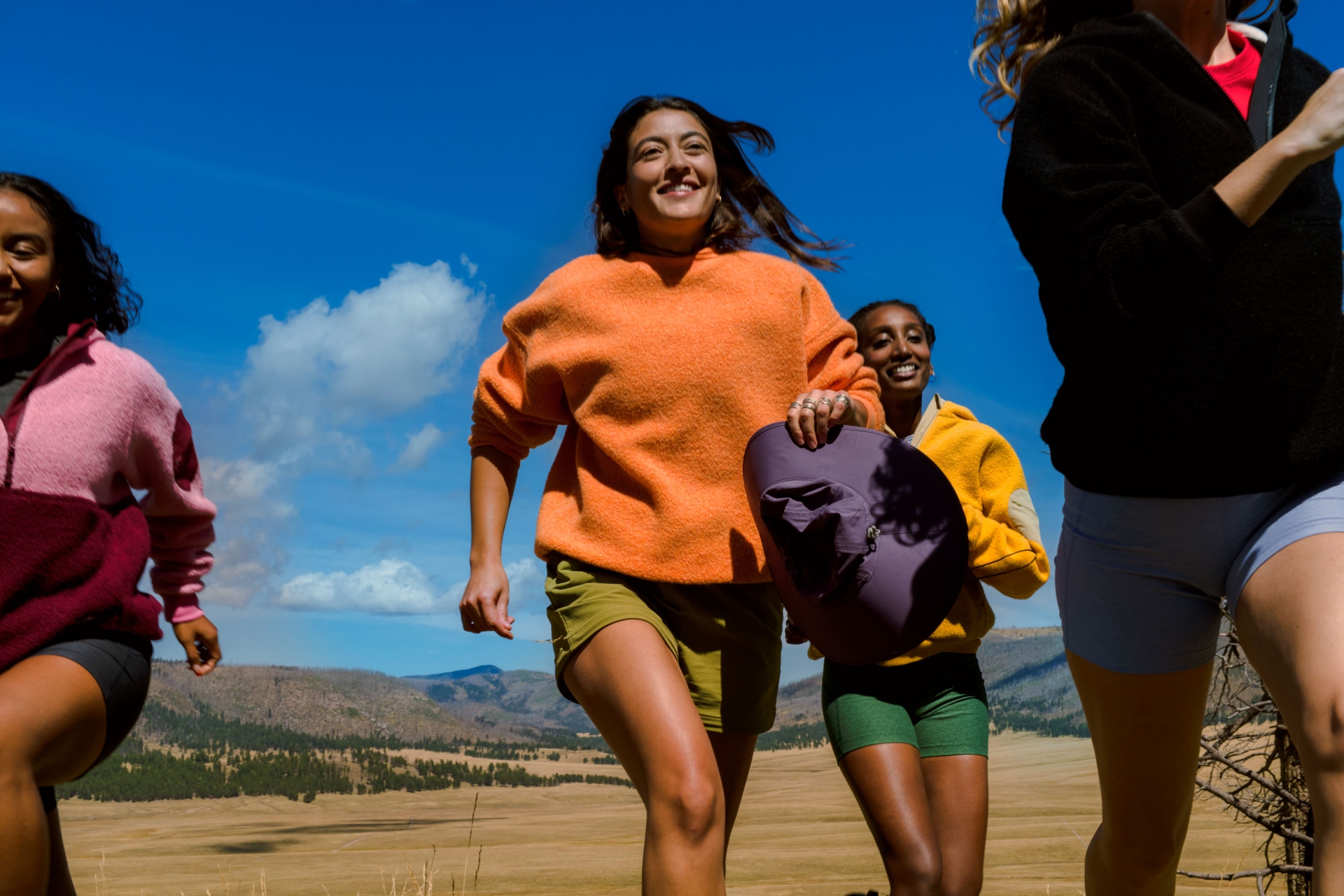 A group of four smiling adult women hike on a grassy hill under a blue sky; landscape view includes trees and distant mountains.