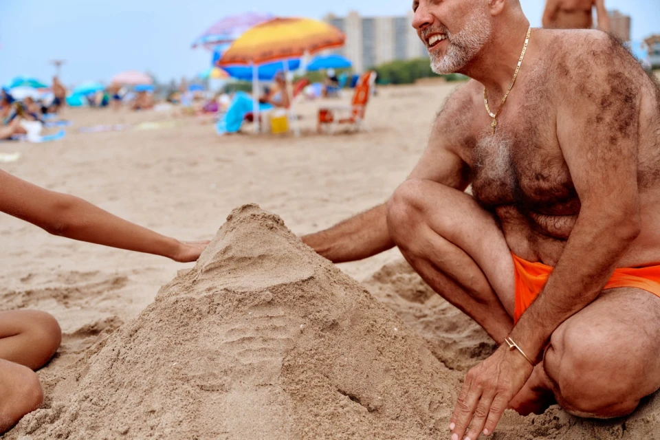 Man and child building a sandcastle on a crowded beach with sun umbrellas and distant buildings under a cloudy sky during the daytime.
