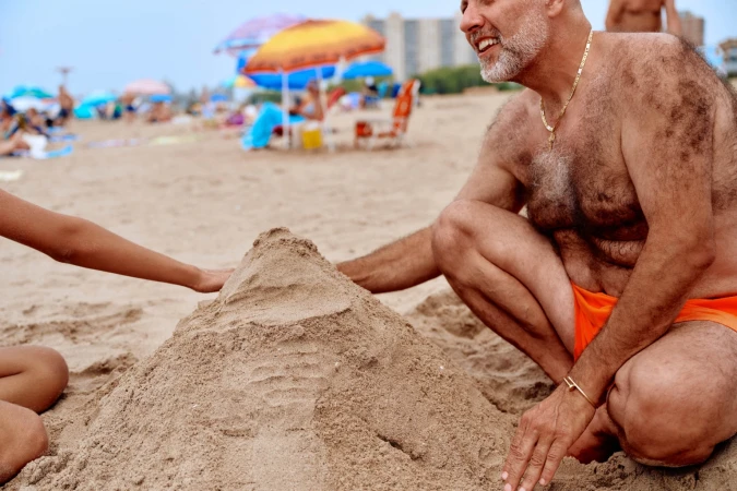 Man and child building a sandcastle on a crowded beach with sun umbrellas and distant buildings under a cloudy sky during the daytime.