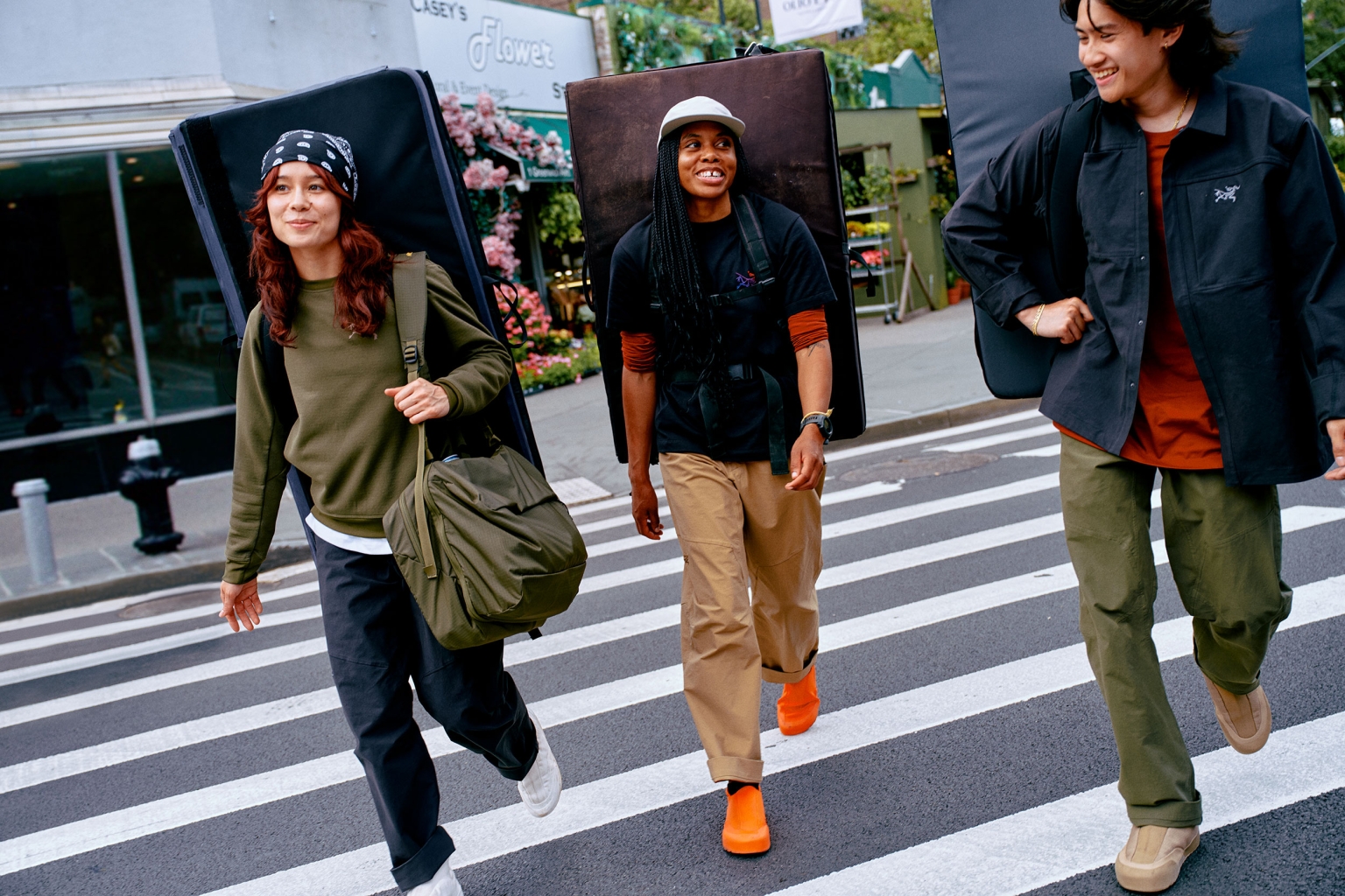 Three people crossing city street in black and olive-green casual outerwear, carrying climbing crash pads and bags, city outdoor background.