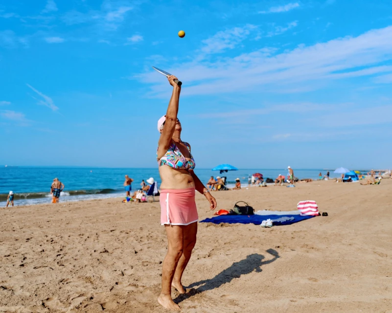 Elderly woman playing paddleball on a sunny beach with other beachgoers. Ocean and blue sky in the background. Active lifestyle.