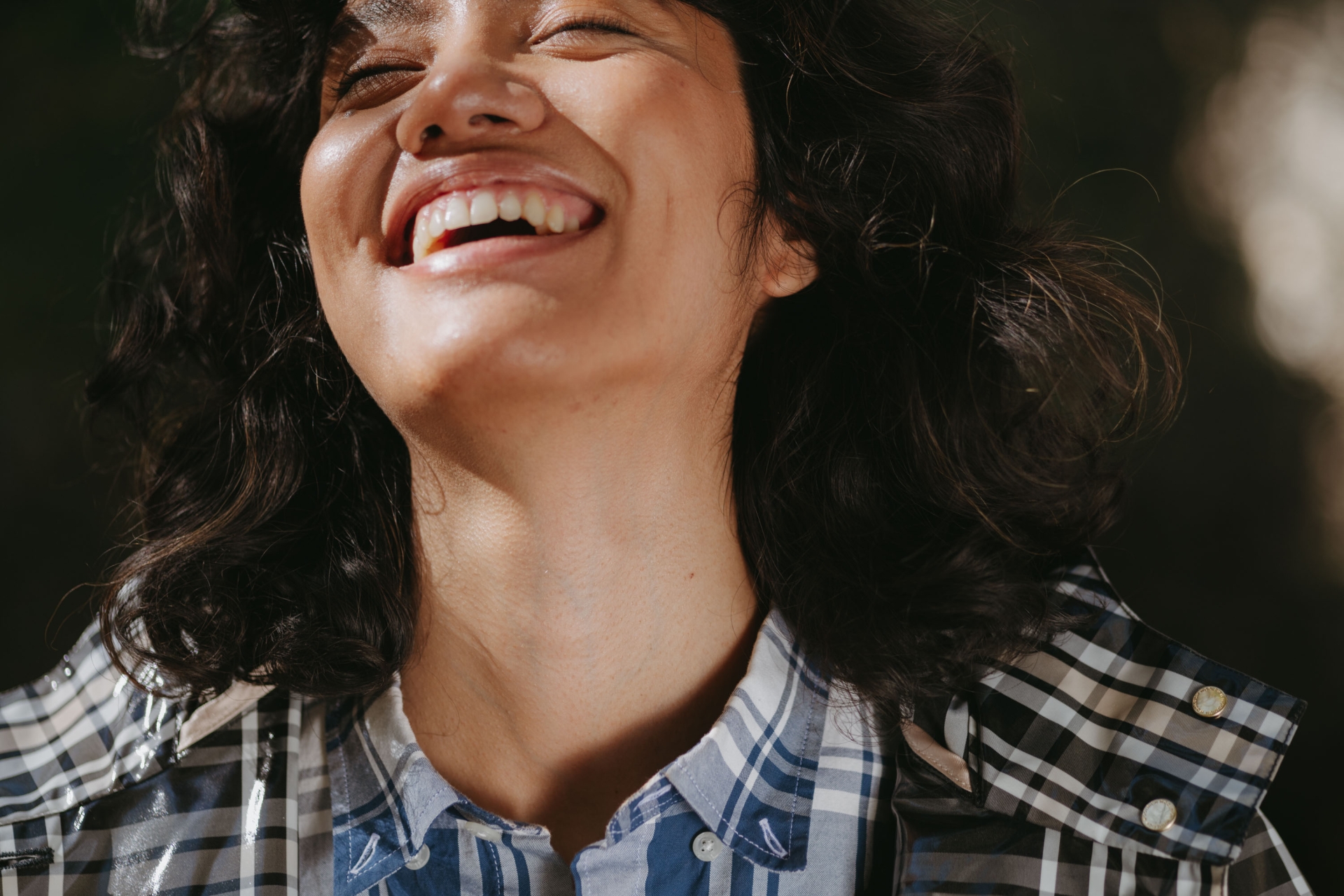 Close-up of a smiling woman with curly dark hair, wearing a plaid shirt with a translucent coat, with an out-of-focus dark background.