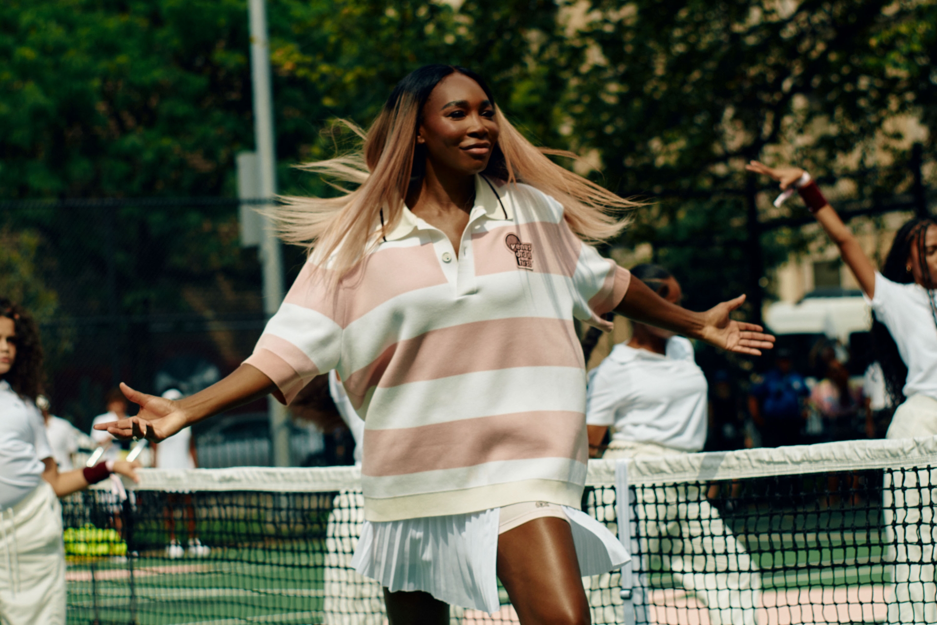 An adult female stands on a tennis court with a group of people behind the net, wearing a striped polo shirt and a white tennis skirt.