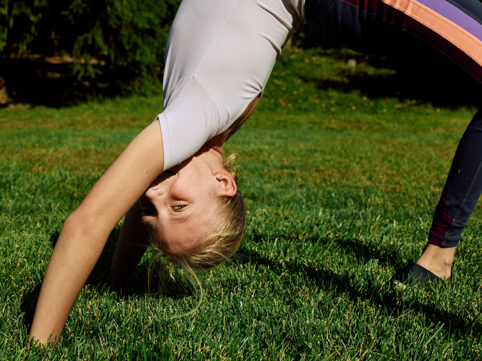 Young woman in downward dog yoga pose outdoors on vibrant green lawn with sunlit trees visible in the background on a sunny day.
